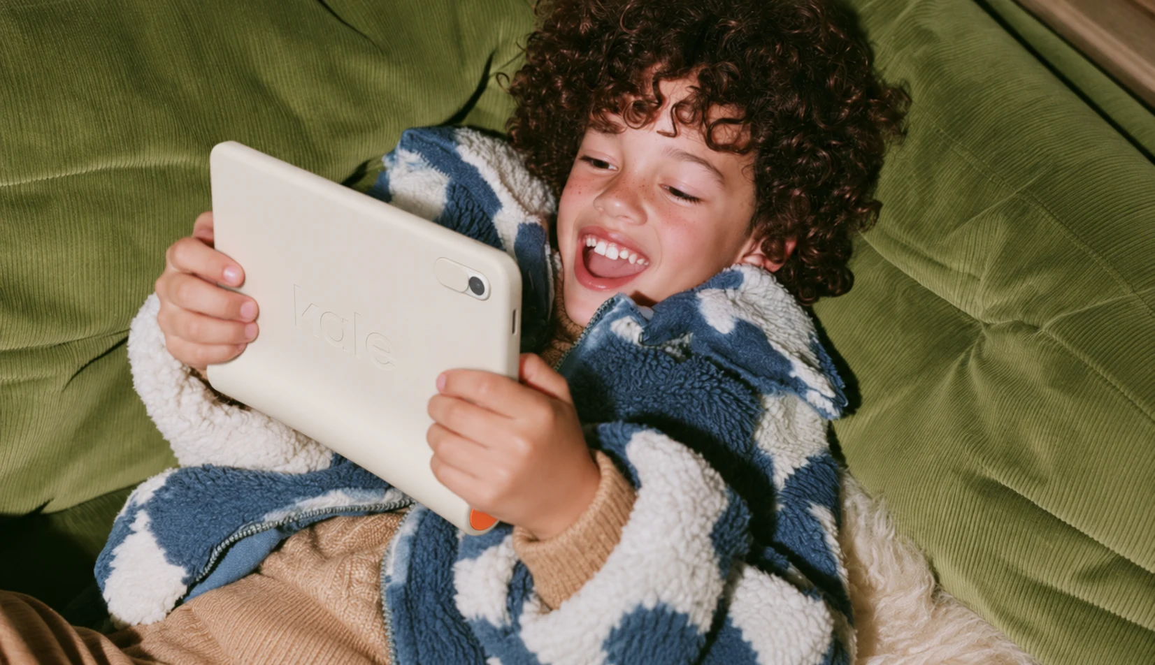 A laughing child in a fuzzy jacket lies on a green couch, holding up Kale to video call with family